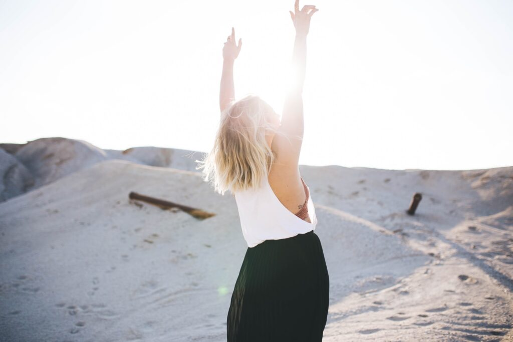 image of a woman wearing white top and black long skirt, with both hands in the air, facing the sun and mounds of sand