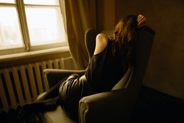 an image of a woman dressed in black sleeveless top and black pants, seated on a chair with her back to the camera, represents the article "If I Was Sick, I was Completely Useless" by Comfortable Hell