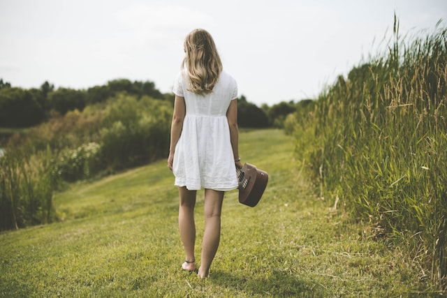 an image of a woman, back to the camera, wearing white dress holding a guitar, walking barefooted on the grass