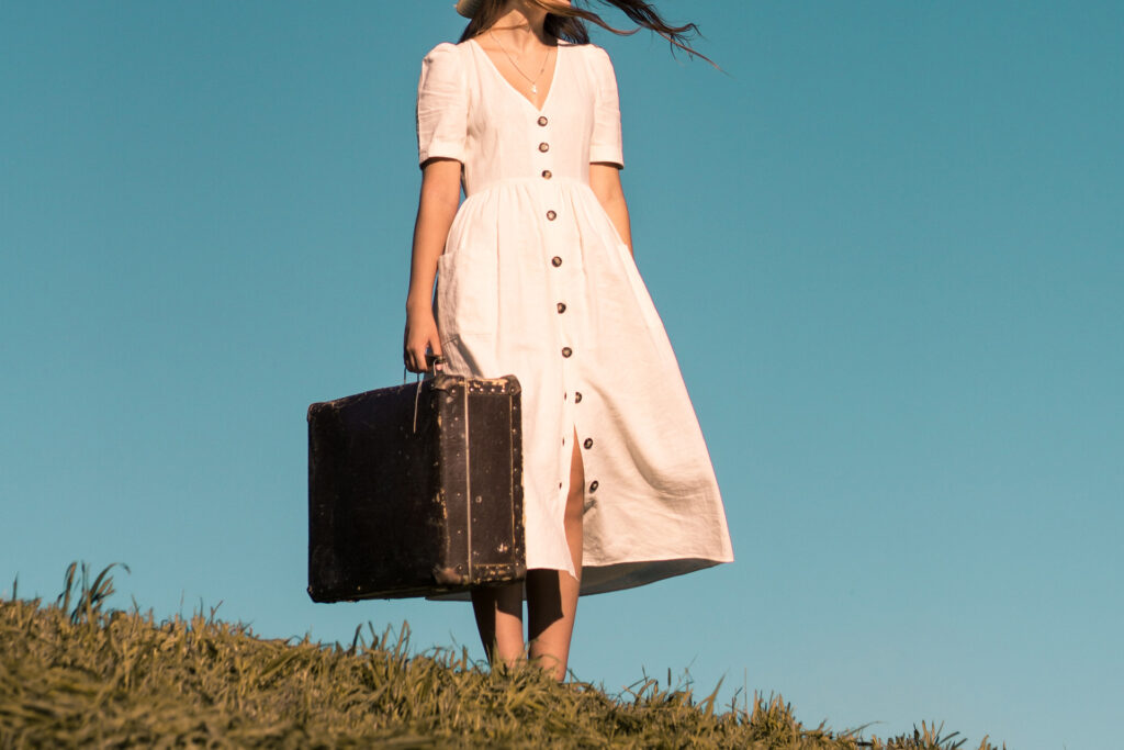 a woman in white dress carrying a vintage suitcase, representing the article "How Do You Feel About You?" by Comfortable Hell